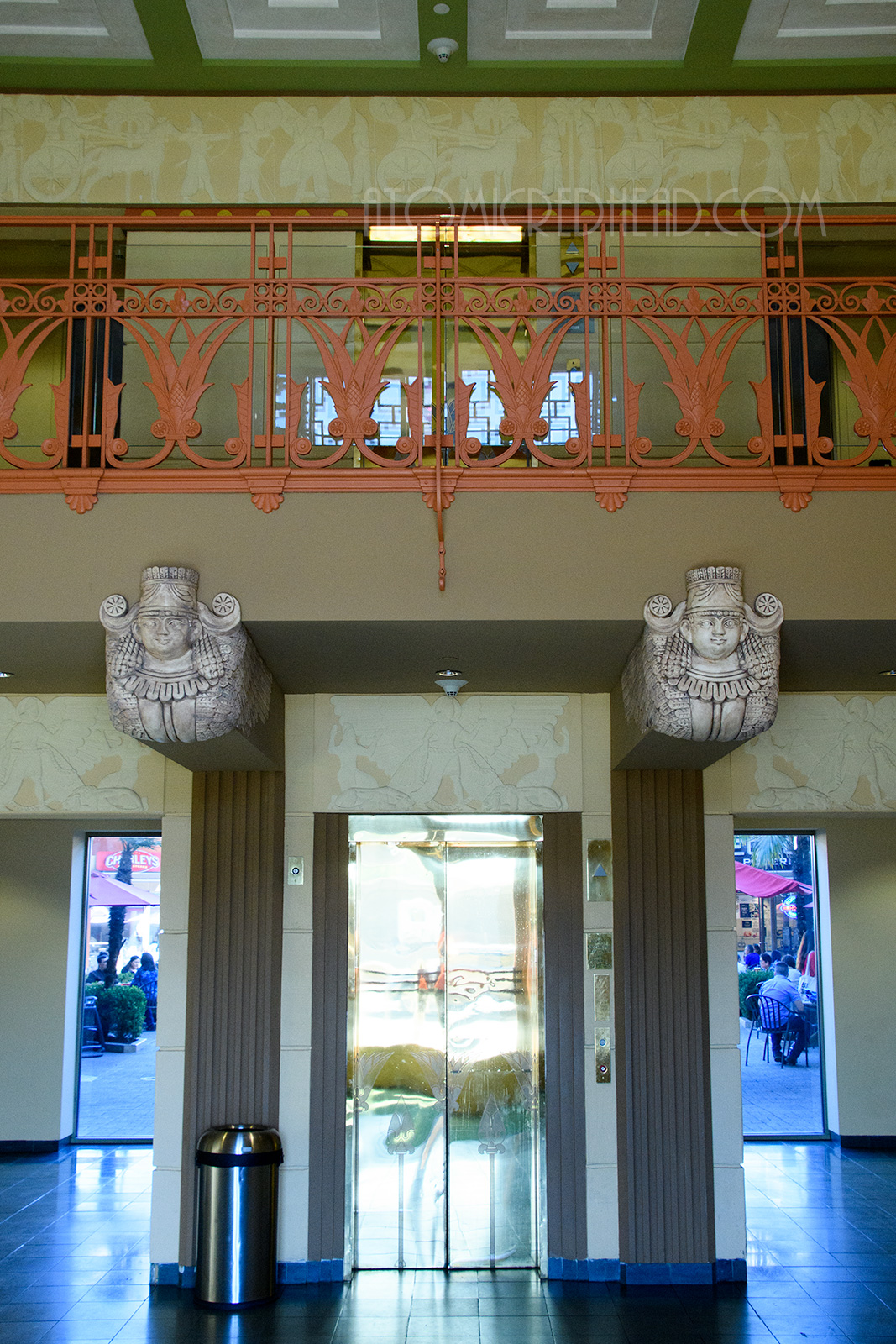 Just inside the building is a central golden elevator, with a balcony above. Holding the balcony are busts of women. The balcony features an iron railing that is painted orange and features lotus flowers.