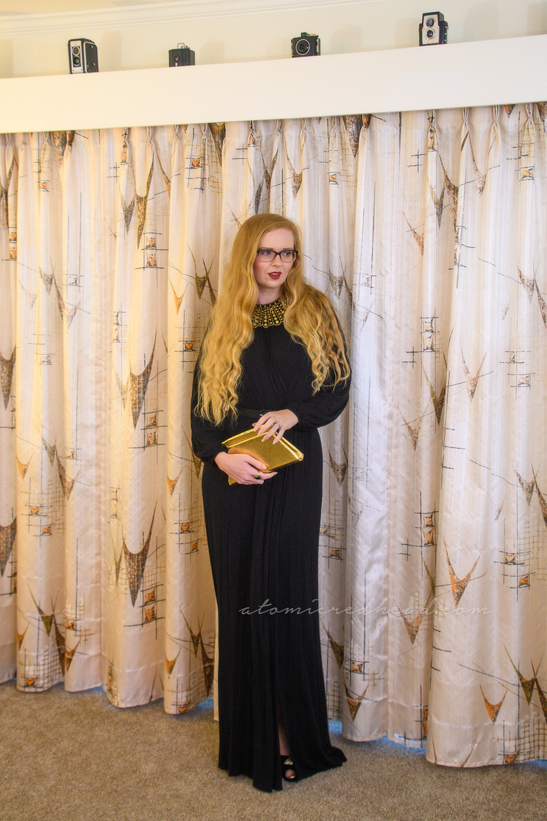 Myself wearing a long sleeve, full length black dress, with a slit up the side, wearing an art decor fan style necklace of gold rhinestones, paired with a gold trapezoid shaped clutch.