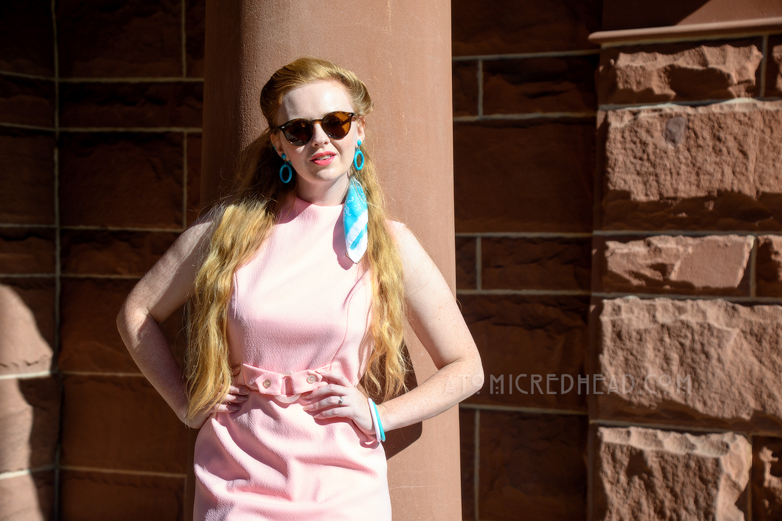 Myself standing in front of the red sandstone building, wearing a sleeveless pink mini dress with a blue scarf in my hair, and blue hoop earrings.
