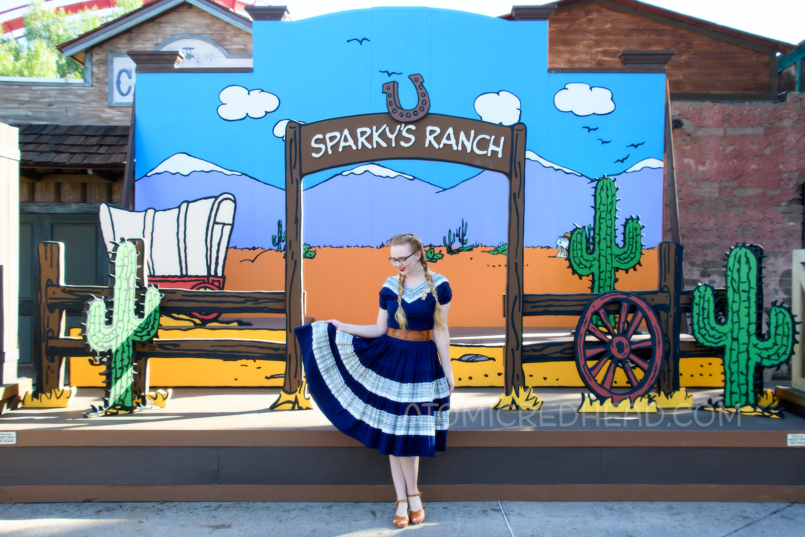 Myself, in front of a Peanuts style backdrop of a desert scene, wearing a navy colored patio set with silver ric-rac trim, and a tooled leather belt, purse, and shoes.
