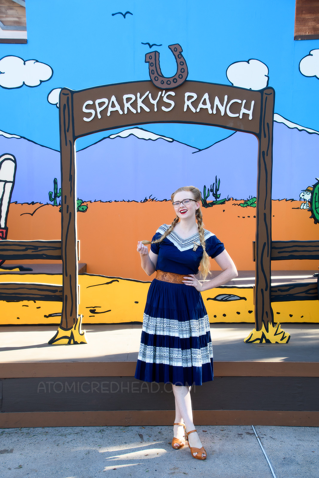 Myself, in front of a Peanuts style backdrop of a desert scene, wearing a navy colored patio set with silver ric-rac trim, and a tooled leather belt, purse, and shoes.