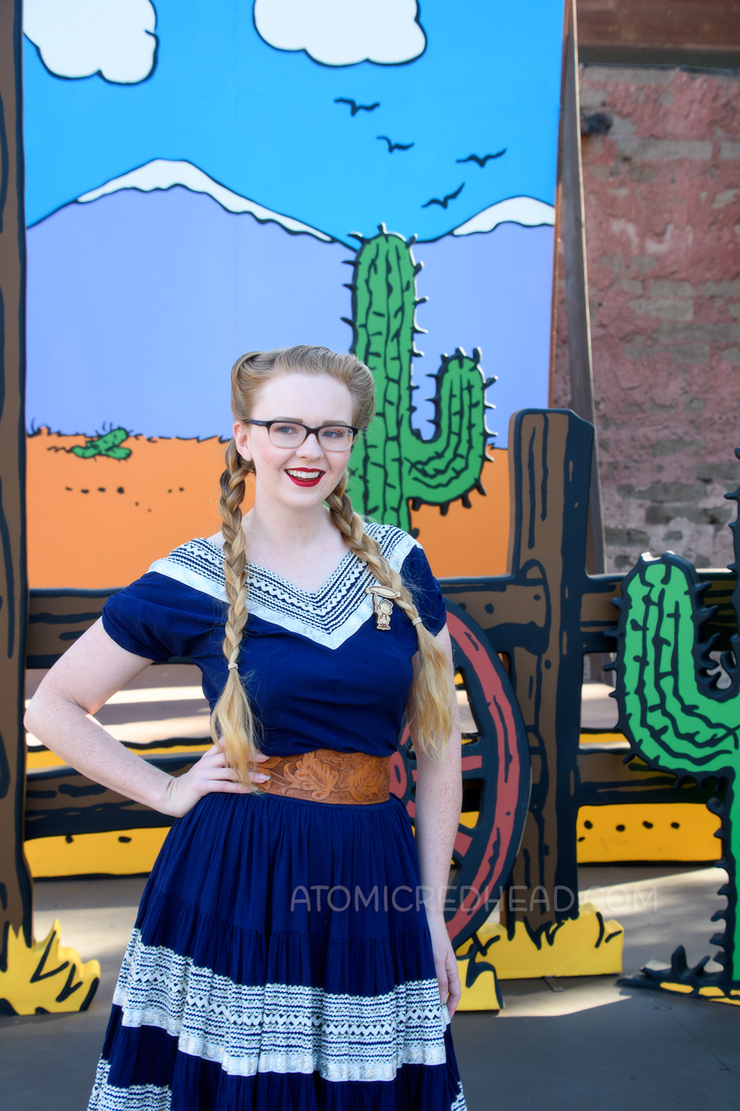 Myself, in front of a Peanuts style backdrop of a desert scene, wearing a navy colored patio set with silver ric-rac trim, and a tooled leather belt, purse, and shoes.