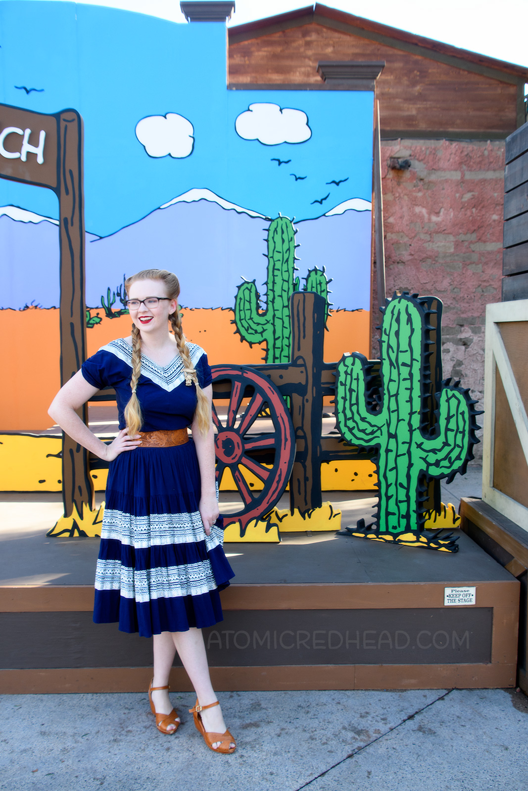 Myself, in front of a Peanuts style backdrop of a desert scene, wearing a navy colored patio set with silver ric-rac trim, and a tooled leather belt, purse, and shoes.