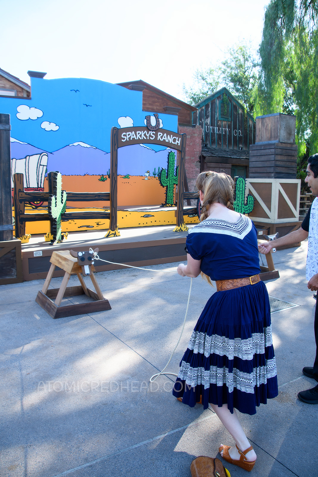 Myself, in front of a Peanuts style backdrop of a desert scene, lassoing a "steer" made from a saw horse, wearing a navy colored patio set with silver ric-rac trim, and a tooled leather belt, purse, and shoes.