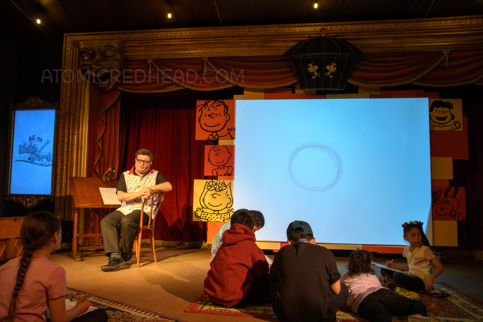 On a stage an artist sits at a desk, behind him a large screen displays what he is drawing on his desk.