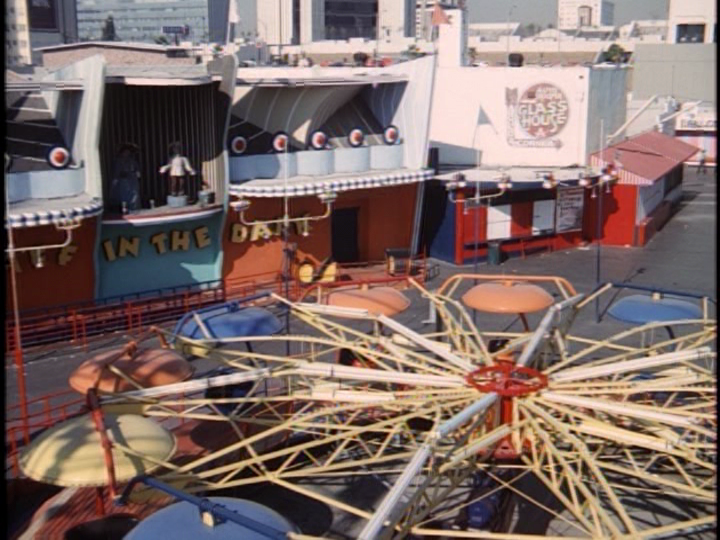 Screencap: A spinning multi-colored ride sits in front of the Laff in the Dark building.