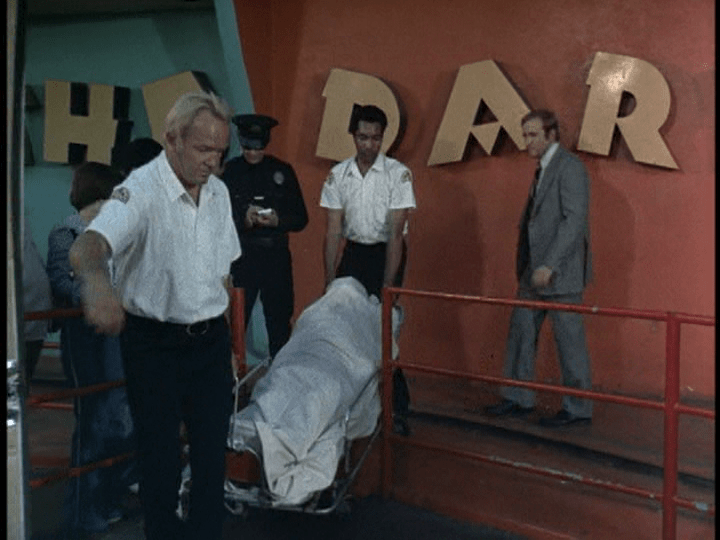Screencap: Men take a body out of the orange and blue Laff in the Dark building, covered in a white sheet.