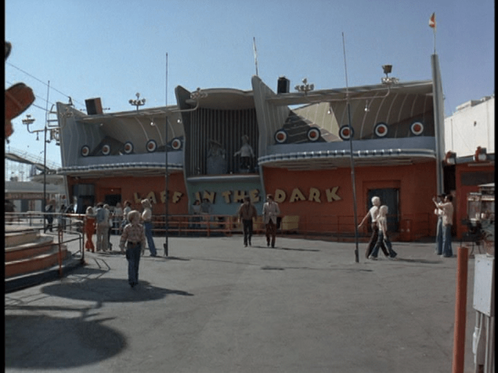 Screencap: A streamline modern looking building painted orange and blue features large yellow letters reading "Laff in the Dark"