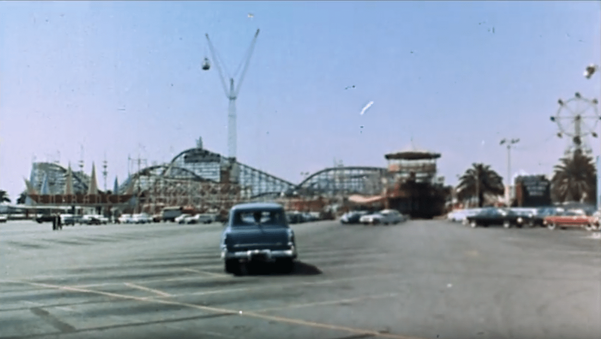 Screencap: A car drives through the parking lot of The Pike, with the tall Cyclone Racer in the background.