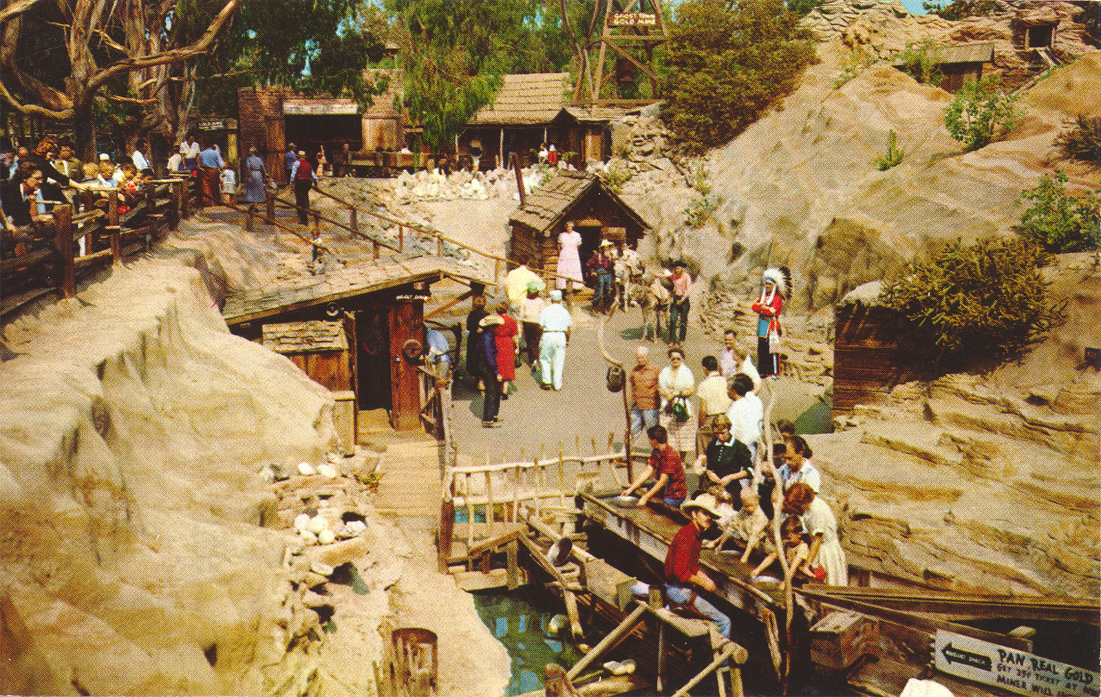 Man made rock formations surround a sluice box where men in wester attire help guests pan for gold.