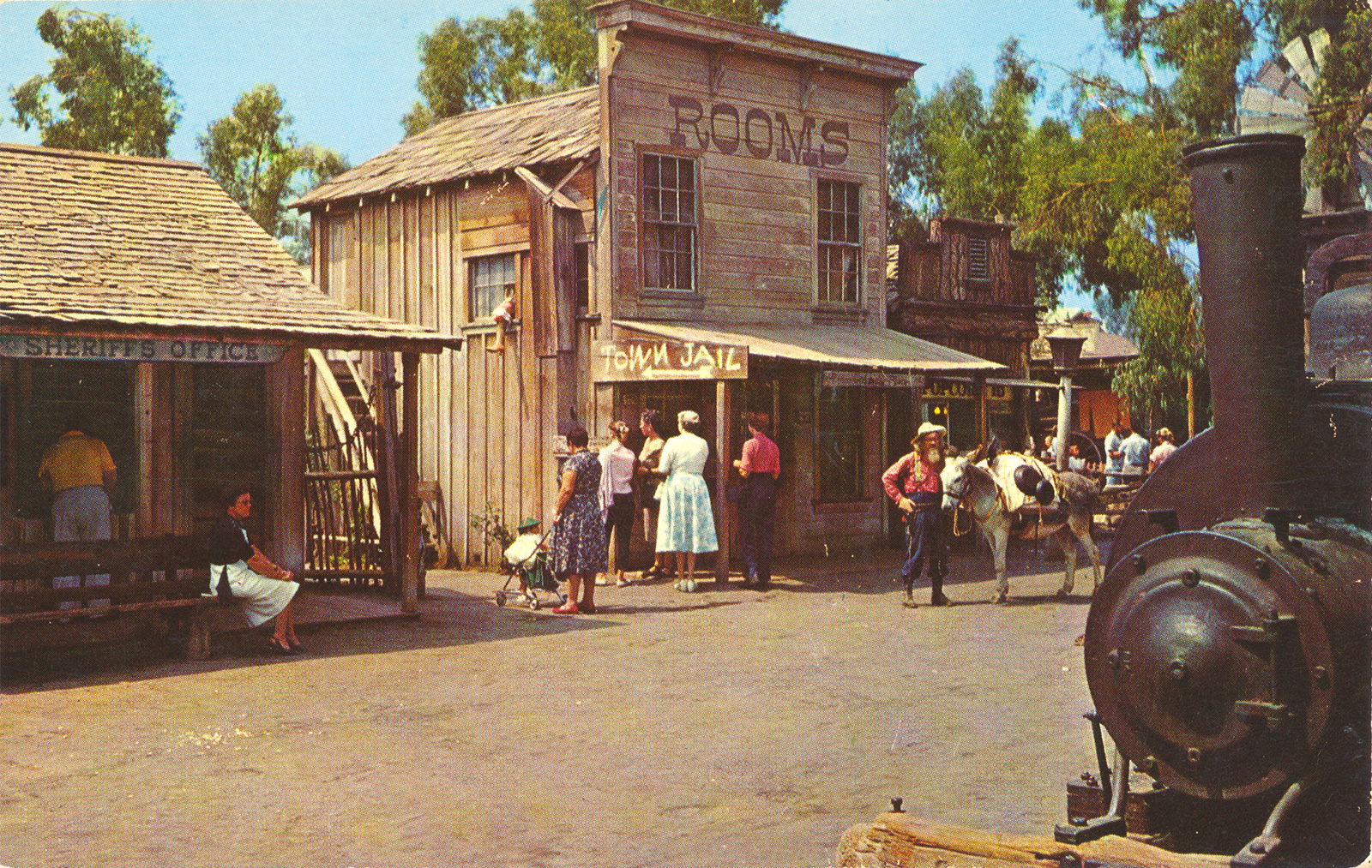 Guests stand near a two story wooden building reading "Rooms" across the top.