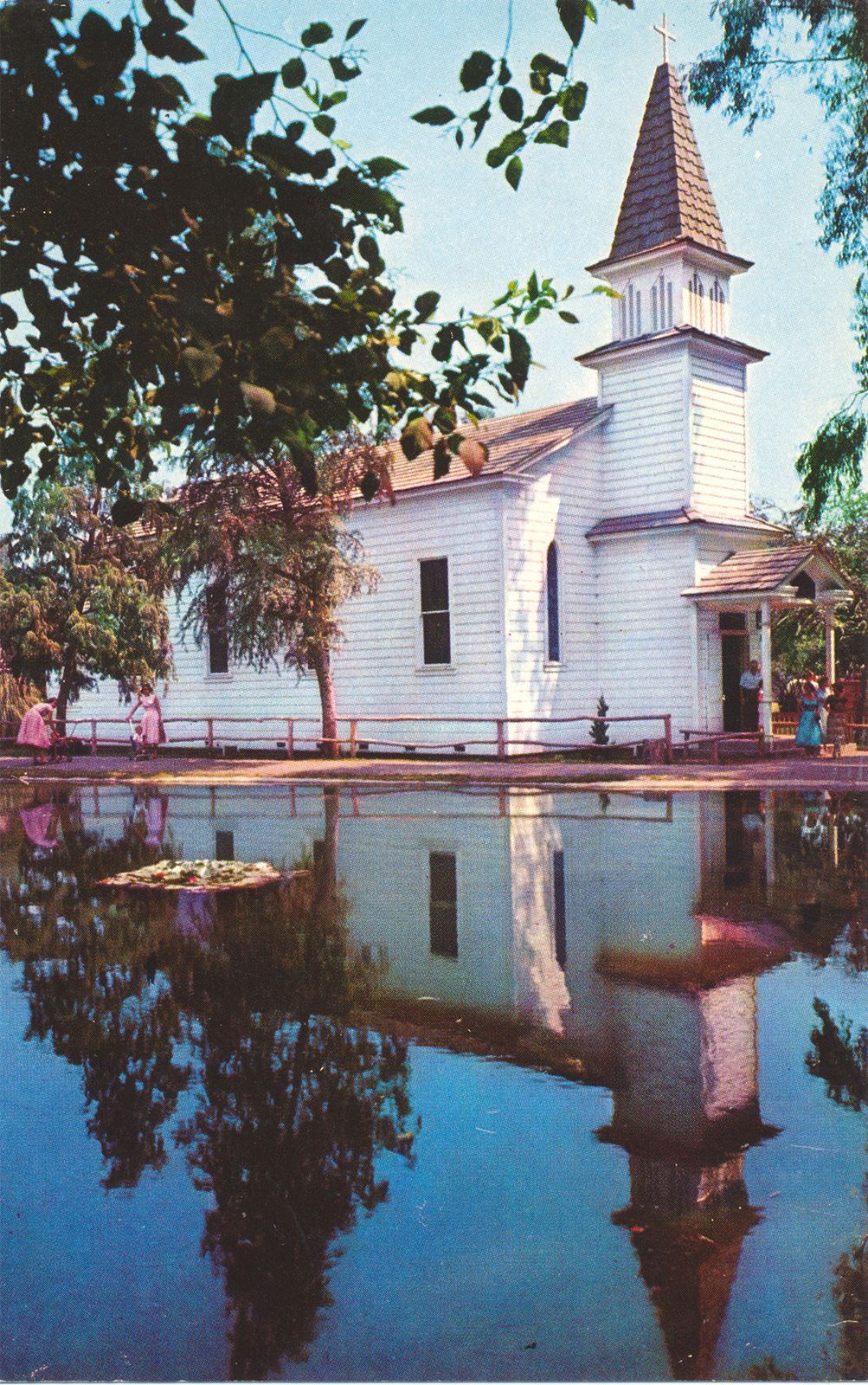 The Church of Reflections, a small classic white church with steeple, sits near a like and has a perfect reflection in the water.