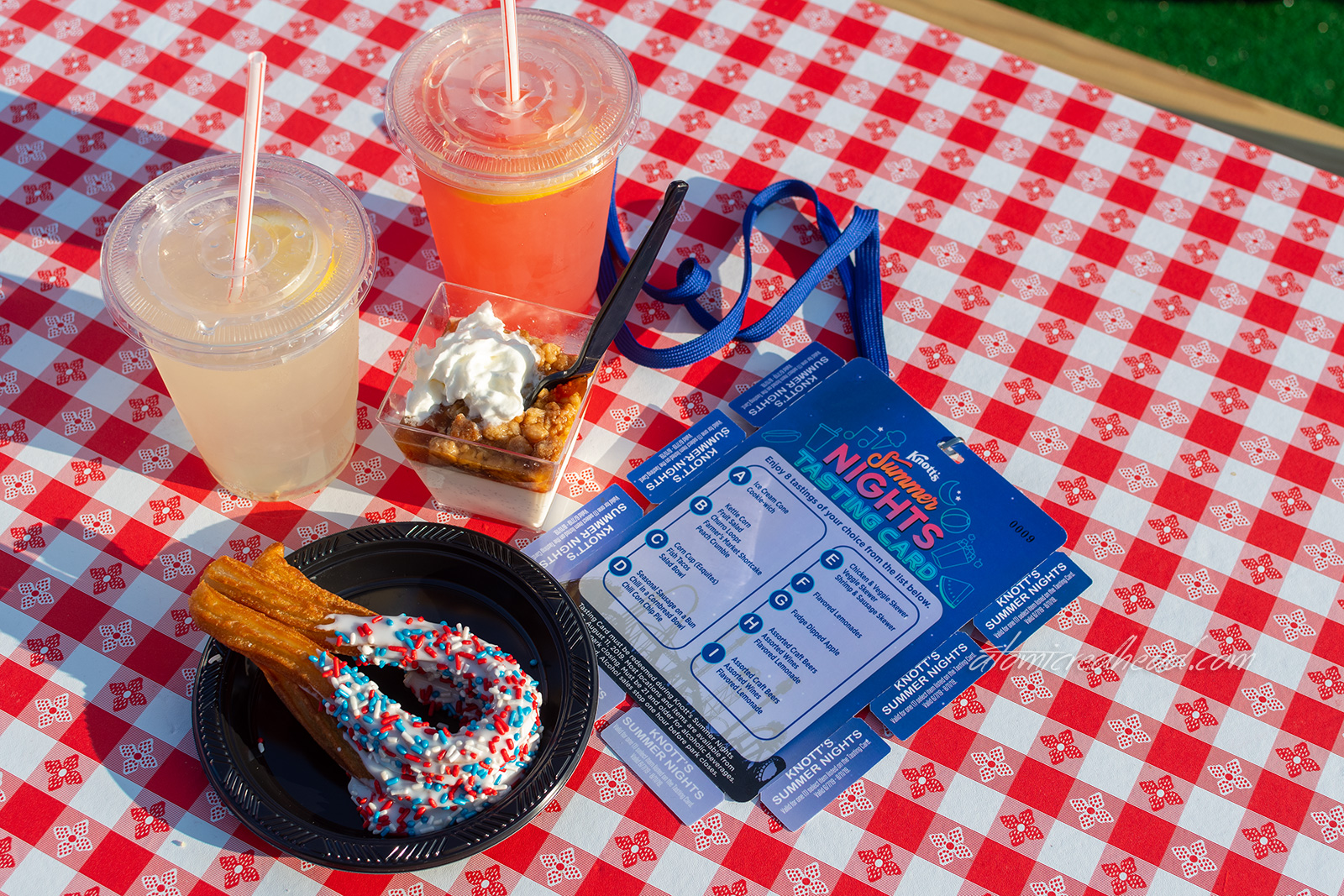 A cup of lemonade, a cup of watermelon lemonade, churros dipped in white frosting with red, white, and blue sprinkles, a small cup with peach cobbler in it, and the blue tasting card sit on a red and white gingham picnic table.