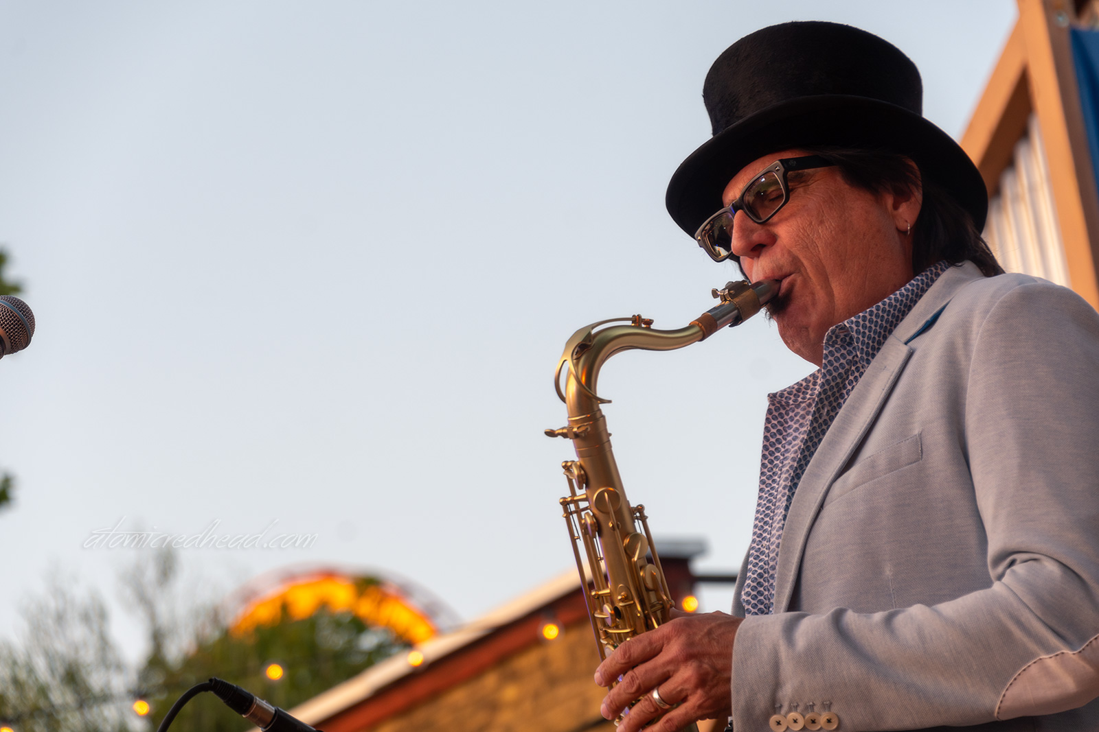 The saxophone player of the zydeco band blows notes while wearing a grey jacket and black top hat.