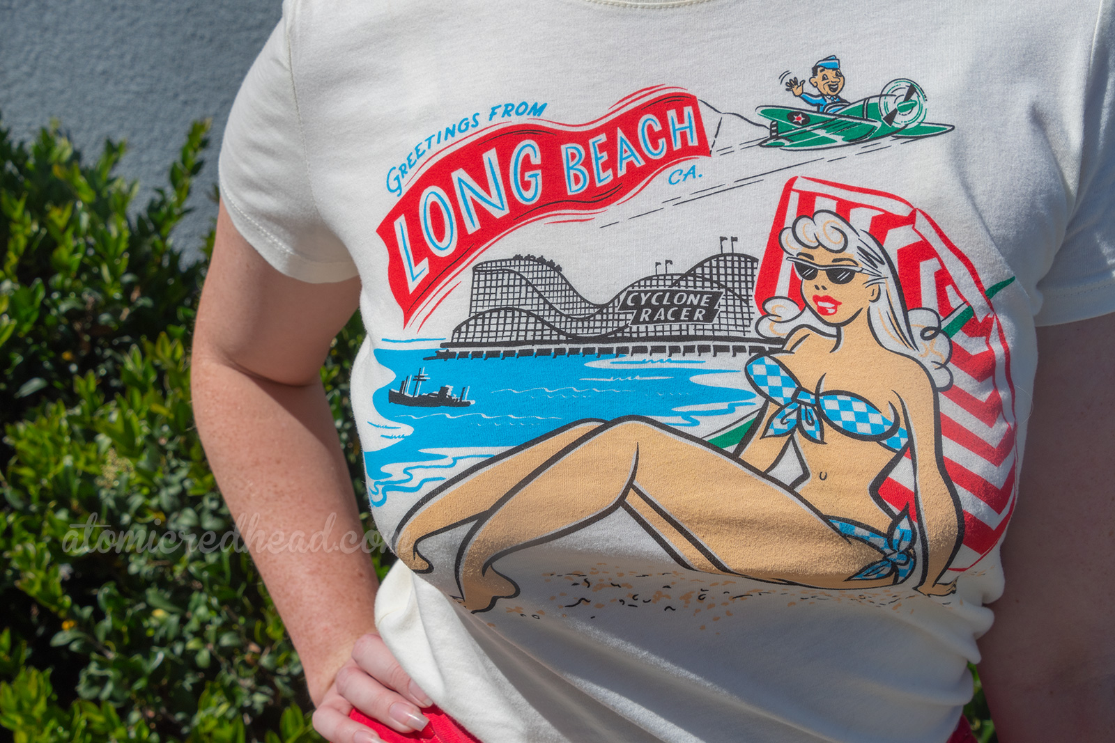 Close up of my t-shirt, a female sunbather wear a blue and white bikini with the ocean and the Cyclone Racer behind her. An airman pilots a green plane with a red banner behind reading "Greetings from Long Beach"