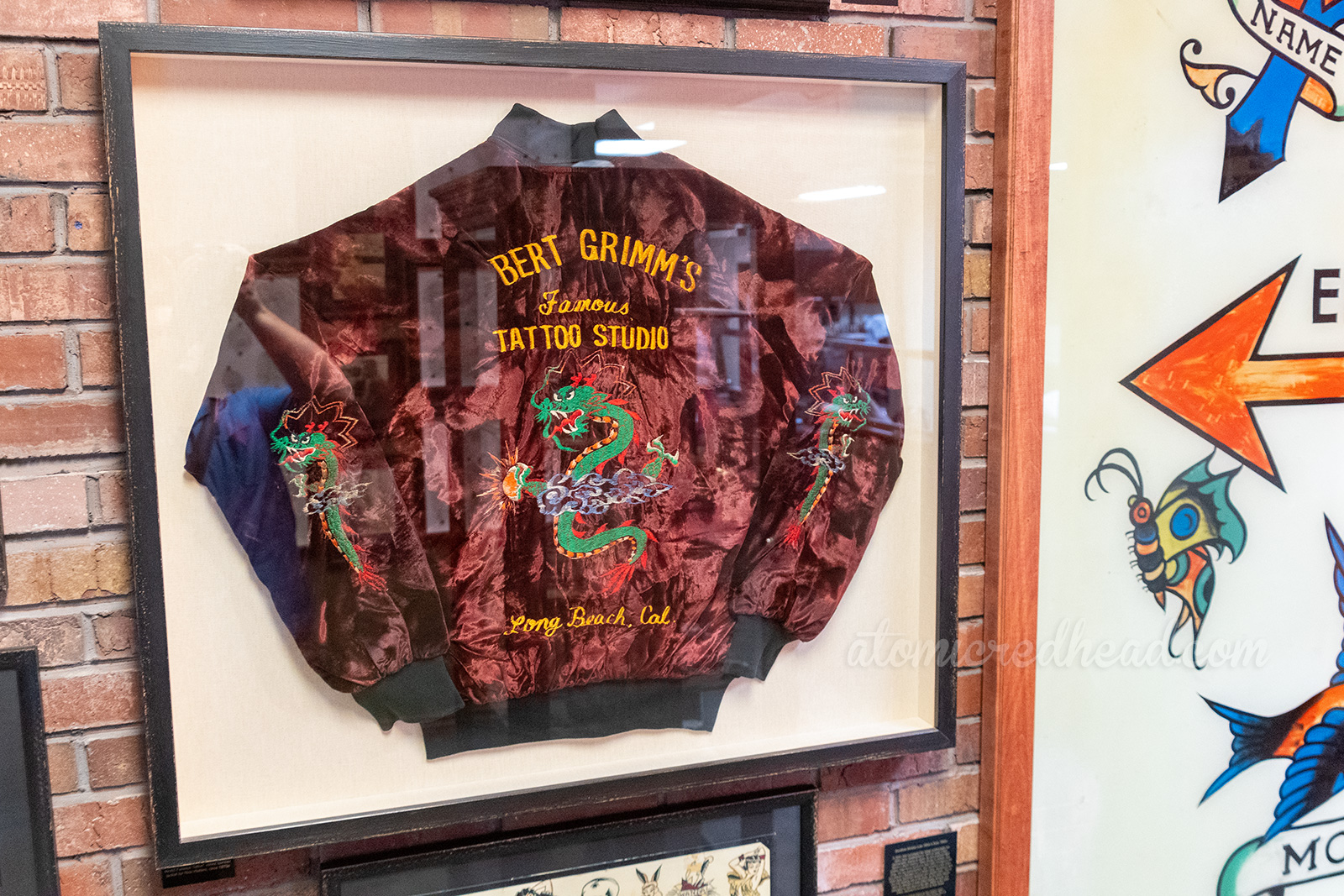 A framed maroon jacket with a Chinese dragon embroidered on the back and sleeves. Yellow lettering reads "Bert Grimm's Famous Tattoo Studio Long Beach, Cal."
