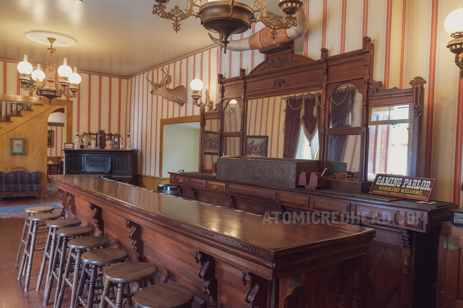 A gloriously dark wood carved bar sits under brass and glass chandeliers. Red and white stripe wallpaper line the walls.