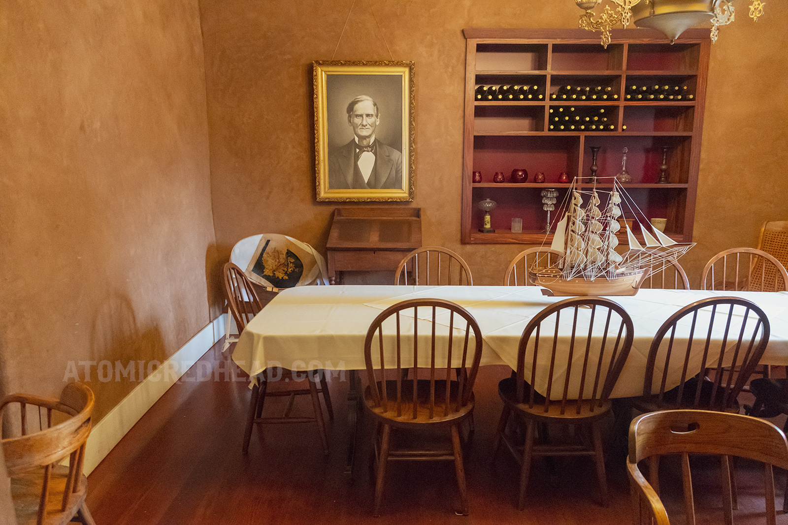 A table inside the saloon featuring a model ship in the center, a black and white portrait of Bandini hangs on the wall.