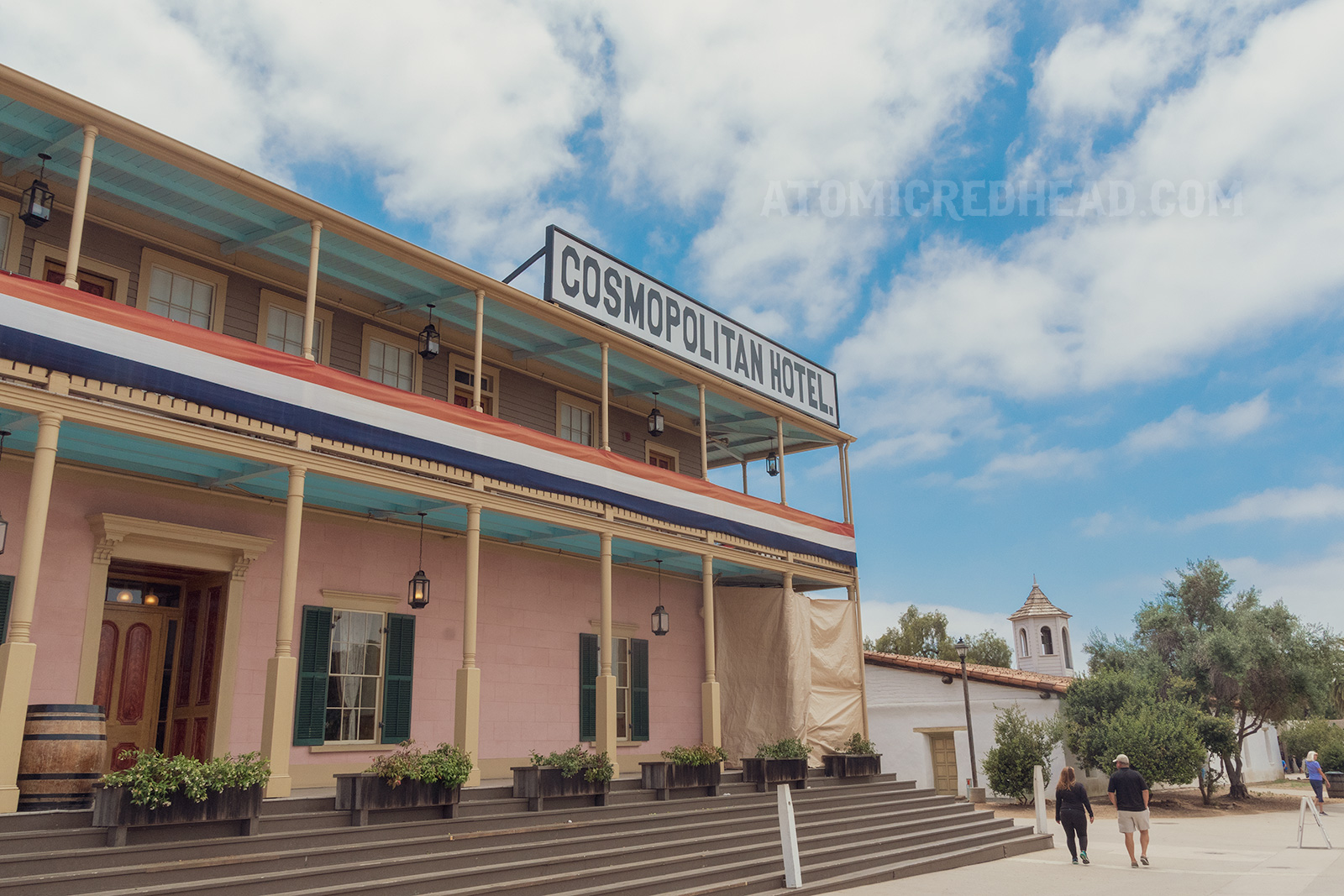 Exterior of the Cosmopolitan, a two story pale pink structure with balcony on the second story. Dark green shutters and pale yellow door trim.