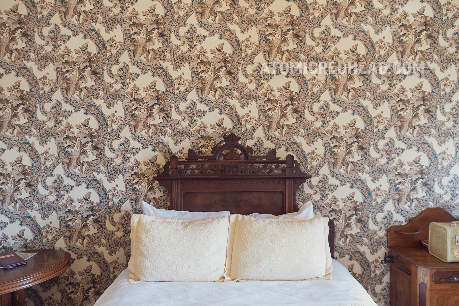 Interior of our room. An antique oak bed sits against a wall. Floral wallpaper in tan, blue, and white wraps around the room.