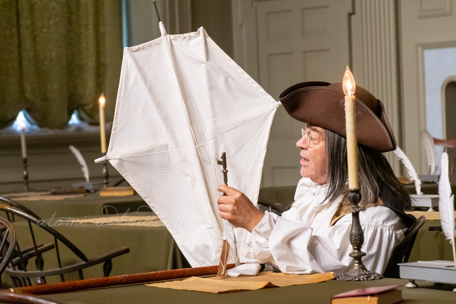 A Ben Franklin reenactor sits at a table holding a kite.