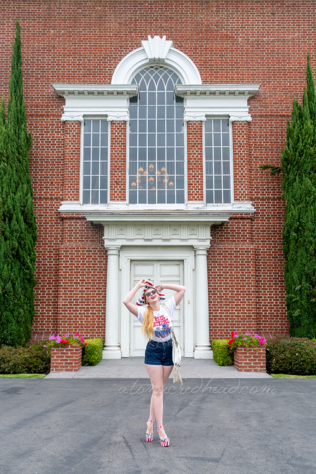 Myself standing outside Independence Hall, a red brick building with white windows and doors, wearing a tricorn hat of red, white and blue stripes, a white t-shirt reading "Paul Revere and the Raiders" with the original flag jutting from the "Raiders" portion, and jean shorts, and platform shoes features red and white stripes and blue with white stars.