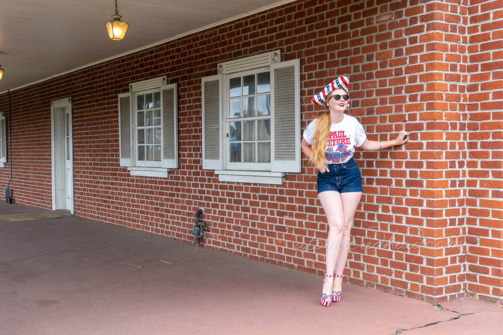 Myself standing outside Independence Hall, a red brick building with white windows and doors, wearing a tricorn hat of red, white and blue stripes, a white t-shirt reading "Paul Revere and the Raiders" with the original flag jutting from the "Raiders" portion, and jean shorts, and platform shoes features red and white stripes and blue with white stars.