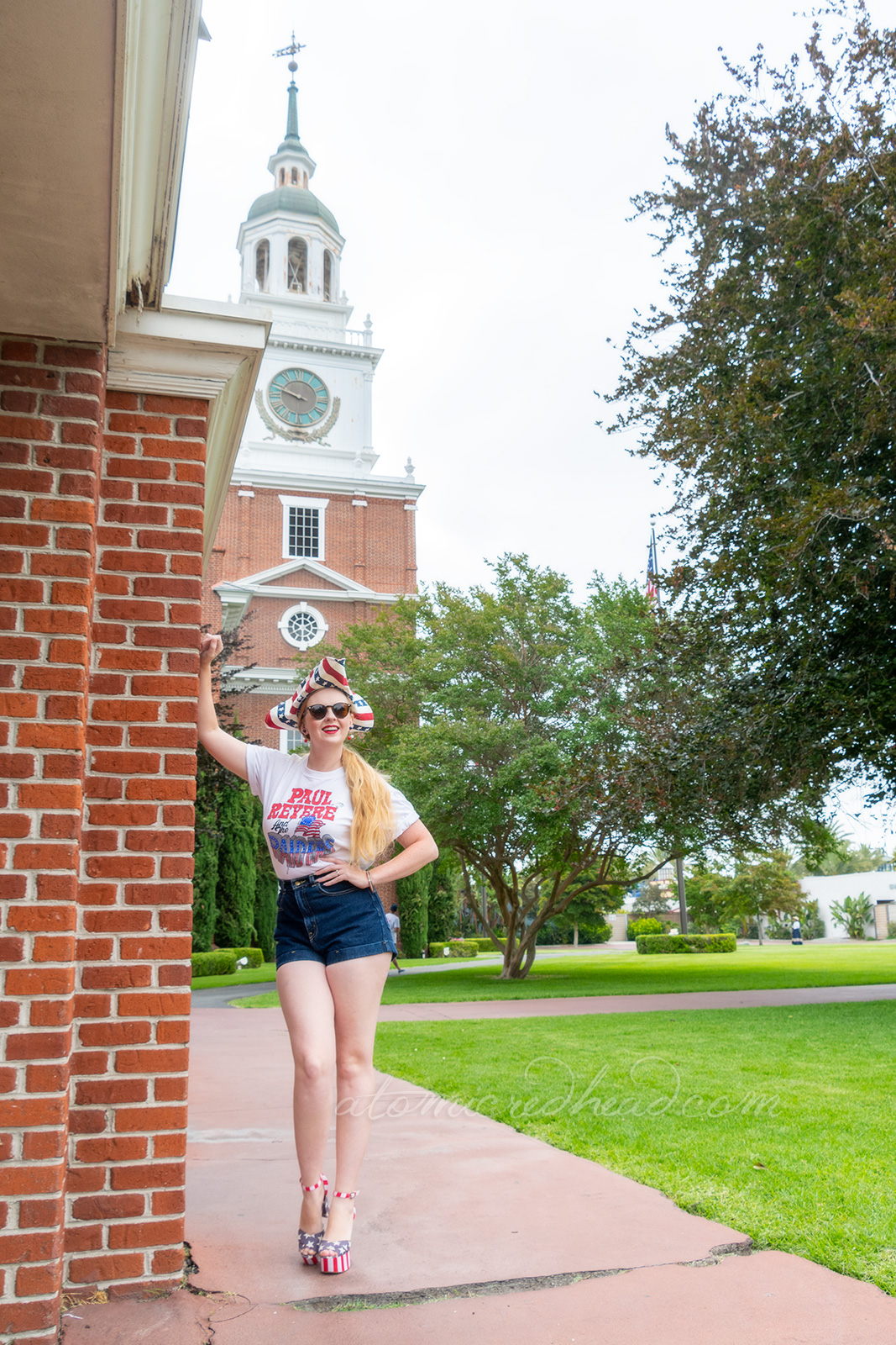 Myself standing outside Independence Hall, a red brick building with white windows and doors, wearing a tricorn hat of red, white and blue stripes, a white t-shirt reading "Paul Revere and the Raiders" with the original flag jutting from the "Raiders" portion, and jean shorts, and platform shoes features red and white stripes and blue with white stars.