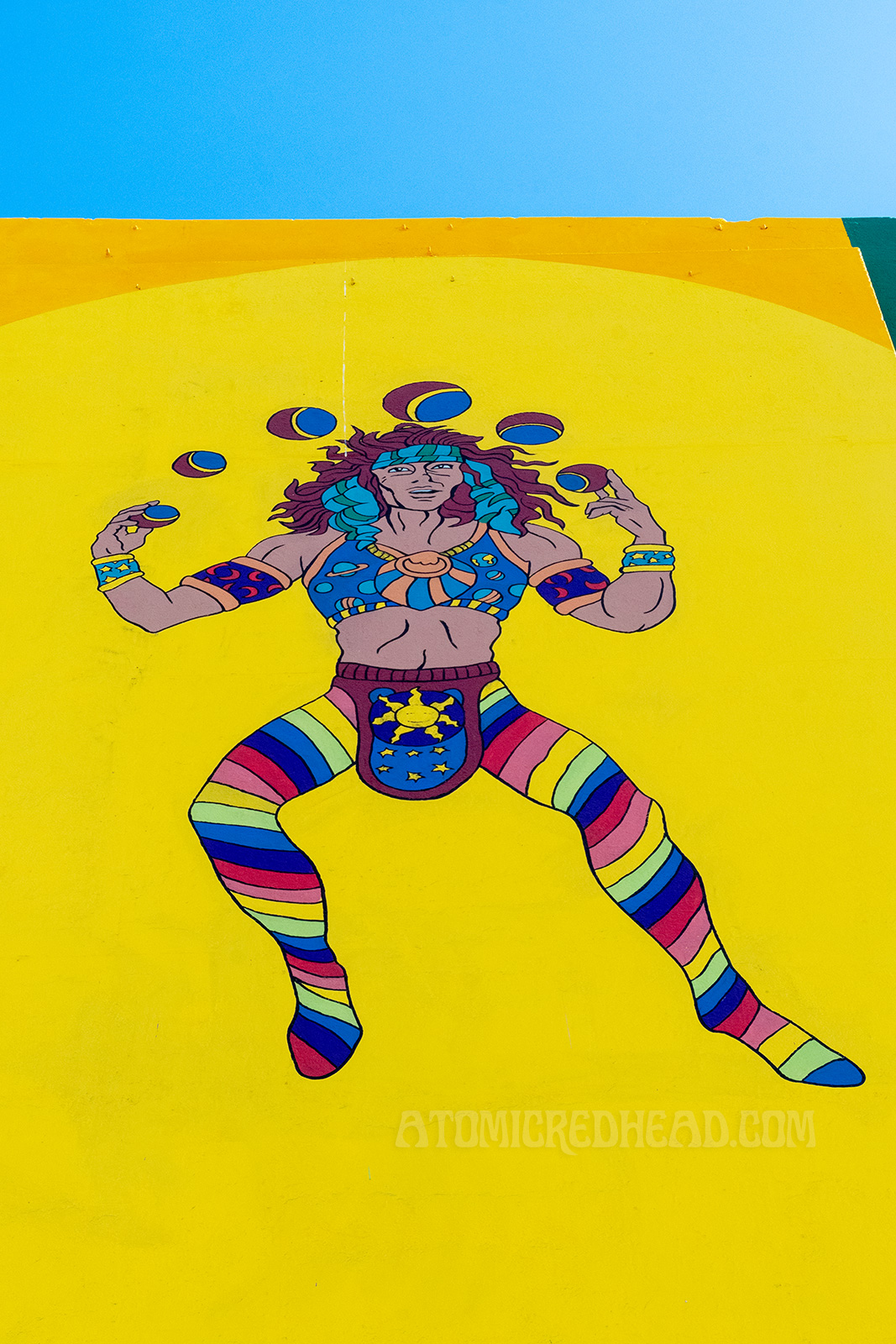 A male juggler with long brown hair and rainbow striped tights, juggles six multi colored balls.