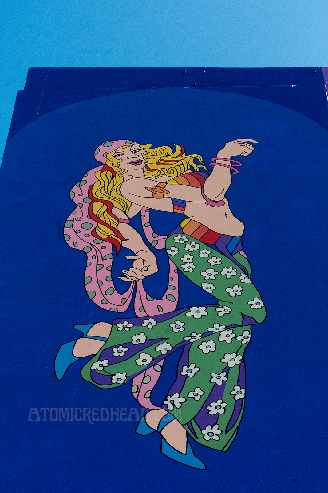 A hippie woman with long blonde hair dances wearing green bell bottoms with white flowers on them.