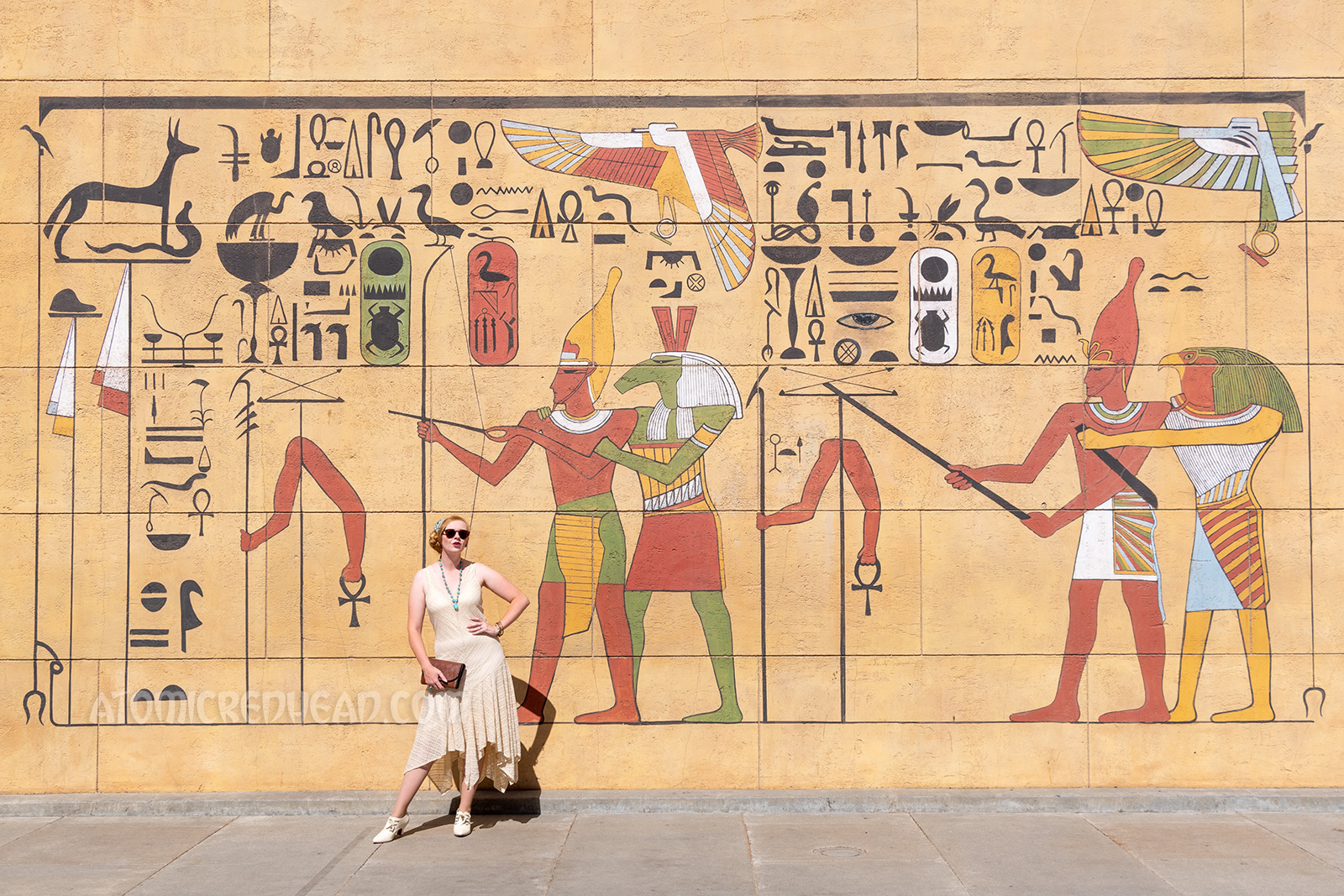 Myself standing in front of a wall in the forecourt that is painted with images of Egyptian gods, kings and hieroglyphs, wearing a sleeveless cream lace dress, a blue scarf with white hieroglyphs, and a blue scarab necklace.