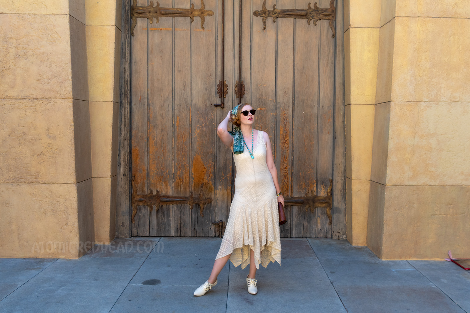 Myself standing in front of a massive pair of wood doors, wearing a sleeveless cream lace dress, a blue scarf with white hieroglyphs, and a blue scarab necklace.