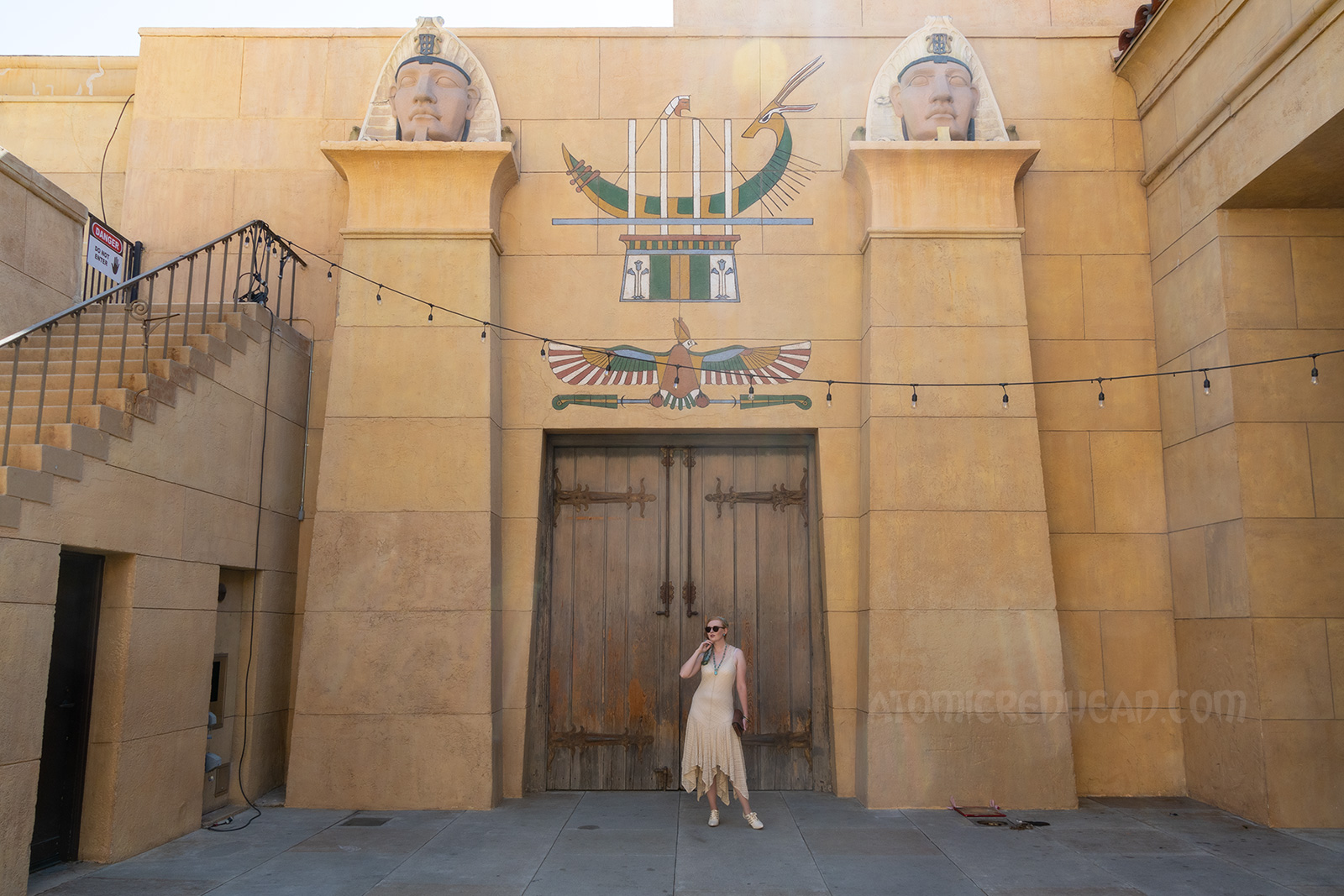 Myself standing in front of a massive pair of wood doors, wearing a sleeveless cream lace dress, a blue scarf with white hieroglyphs, and a blue scarab necklace. Above the two doors of painted hieroglyphs 