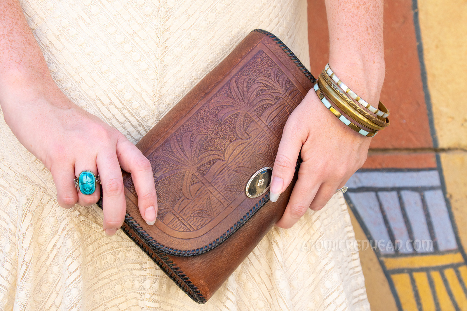 Close-up of my purse and jewelry. Purse is brown leather with an image of palm trees. Gold tone bracelets, on one wrist, a blue scarab ring on the other hang.