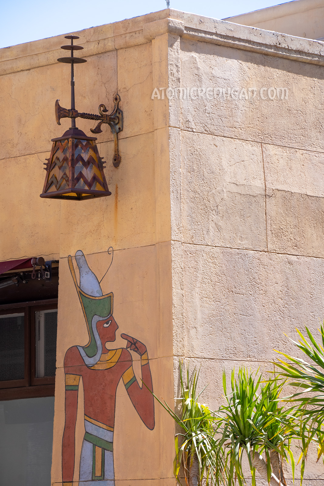 A unique Egyptian influenced lamp, with an amber colored glass shade, and iron zig-zag and lotus flower design hangs above a mural of an Egyptian man.