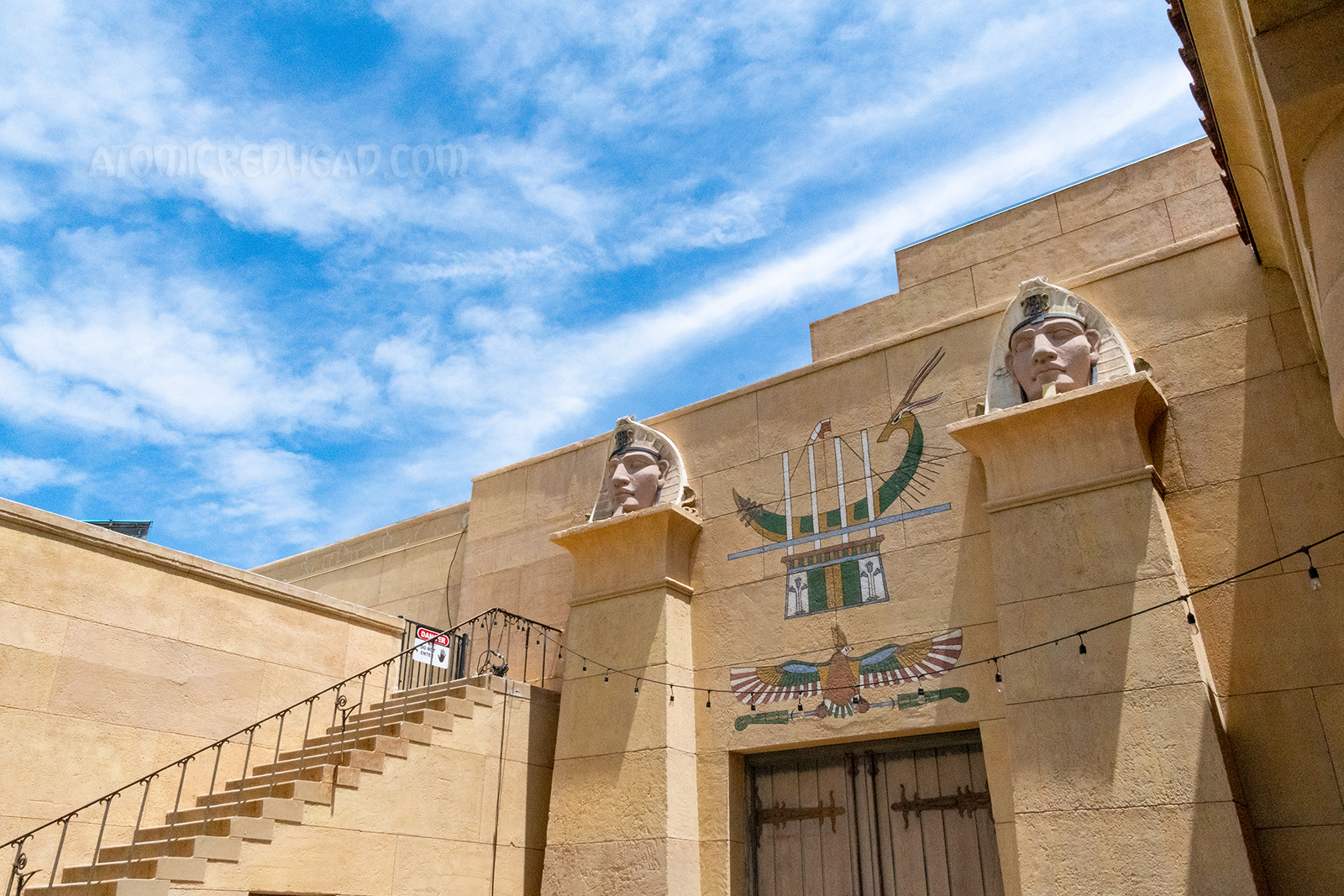 A pair fo wooden doors sit between two pillars, above the doors is a mural of a hawk and a boat, atop the pillars are images of Egyptian pharaohs.