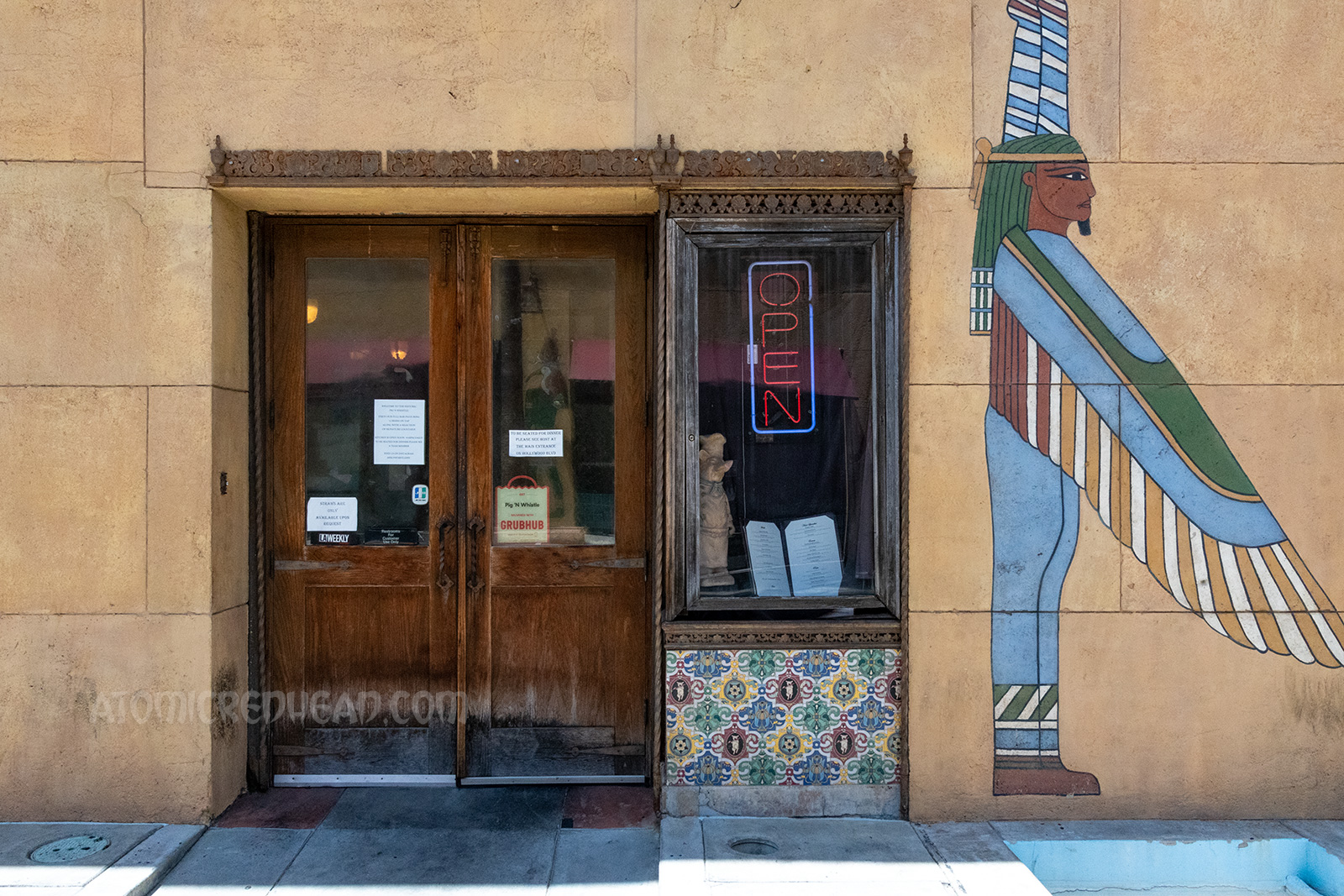 A wooden door and window with tile work below it enters into the neighboring restaurant of Pig n Whistle. Painted to the right is a mural of winged Egyptian gods.