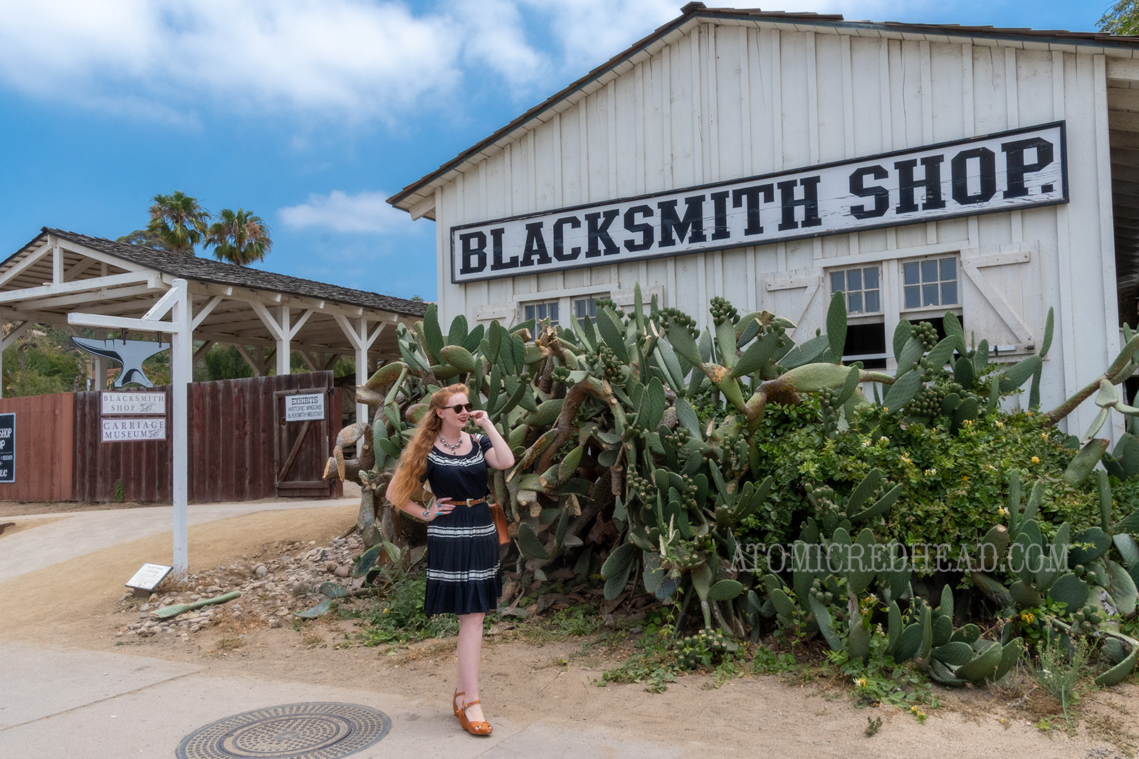 Myself standing in front of the blacksmith shop, where a giant prickly pear cactus resides, wearing a black dress with silver trim.