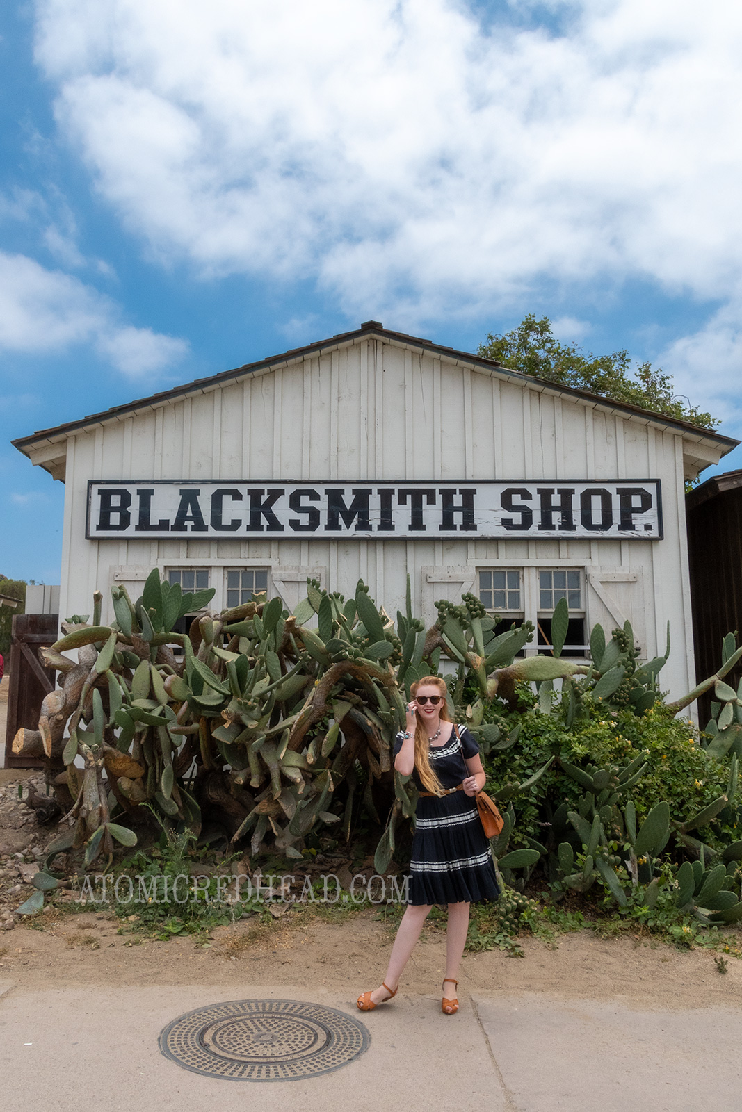 Myself standing in front of the blacksmith shop, where a giant prickly pear cactus resides, wearing a black dress with silver trim.