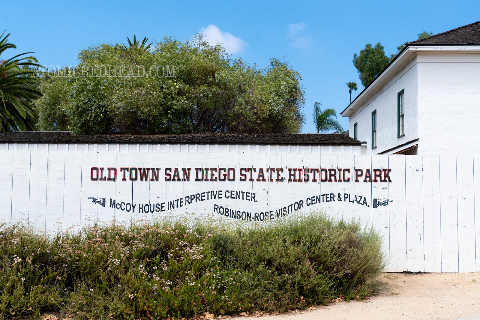 A white fence with black text reads "Old Town San Diego State Historic Park" below in wavy black letters and painted hands pointing reads "McCoy House Interpretive Center" one way and "Robinson Rose Visitor Center & Plaza" the other way.