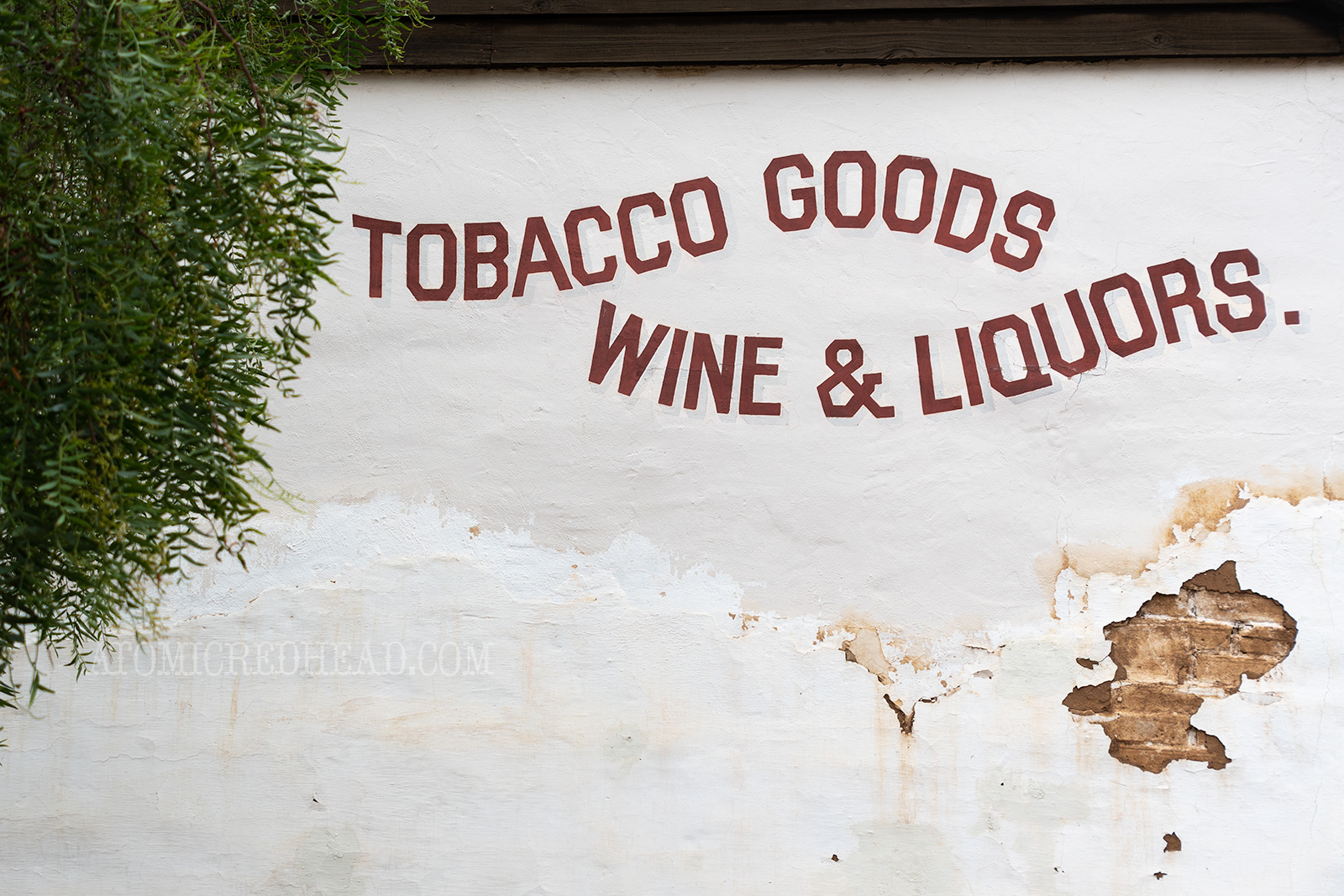 An old adobe wall features waving text reading "Tobacco Goods Wine & Liquor"
