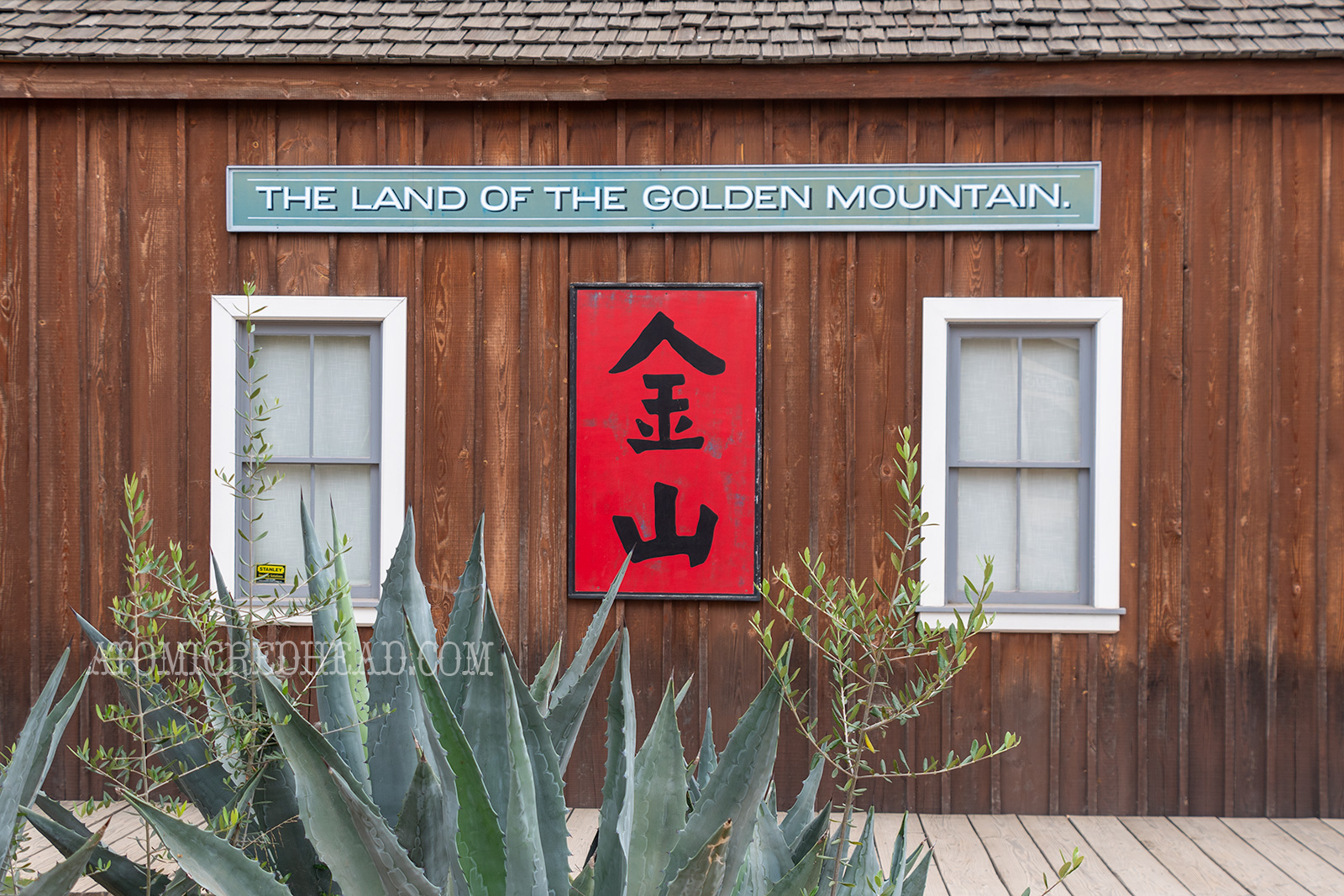 Exterior of an unpainted wood building. A green sign reads "The Land of the Golden Mountain" a red sign below features Chinese characters. 