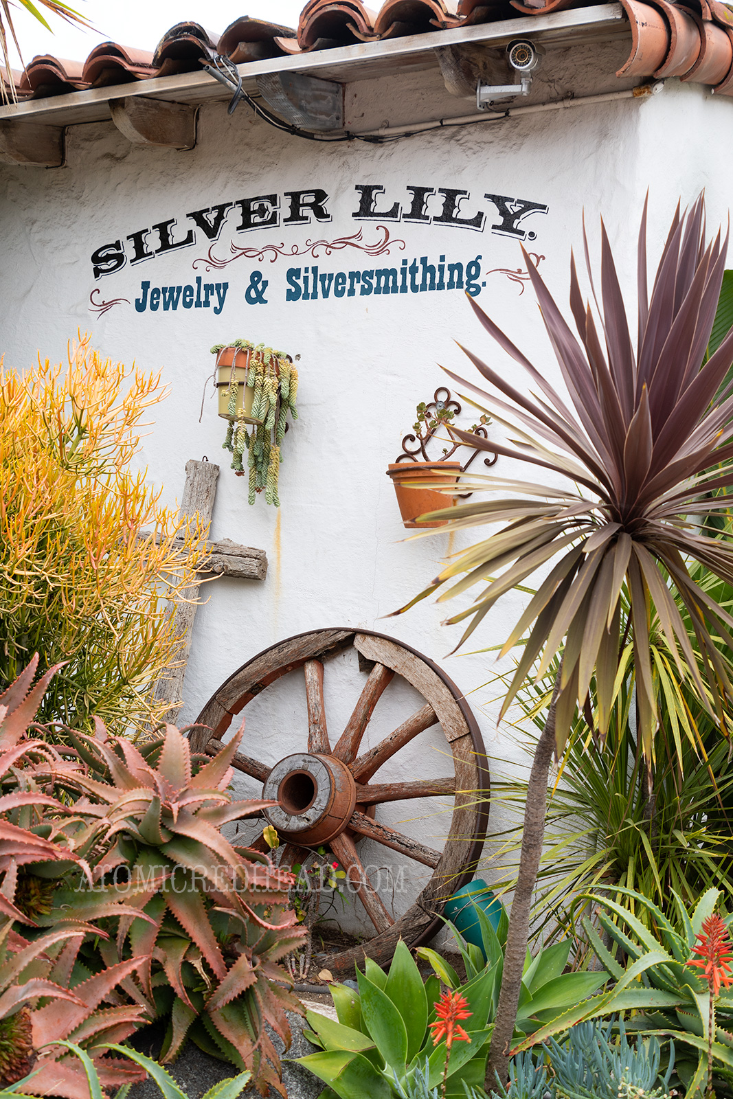 A white adobe wall reads "Silver Lily Jewelry & Silversmithing" with succulents and cacti below, along with an old wagon wheel.
