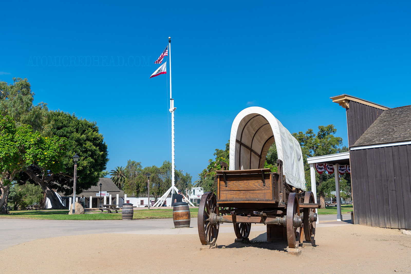 A covered wagon sits near the green lawn of the plaza with a tall flag pole features the American and California flags.
