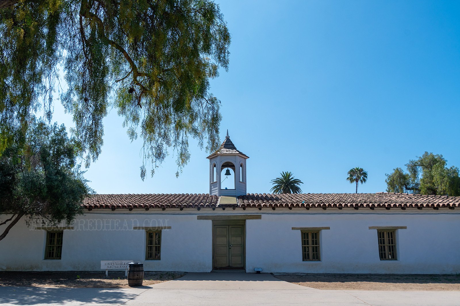 A single story adobe home, with a cupola atop in the middle, and red tile roof and green doors and shutters. 