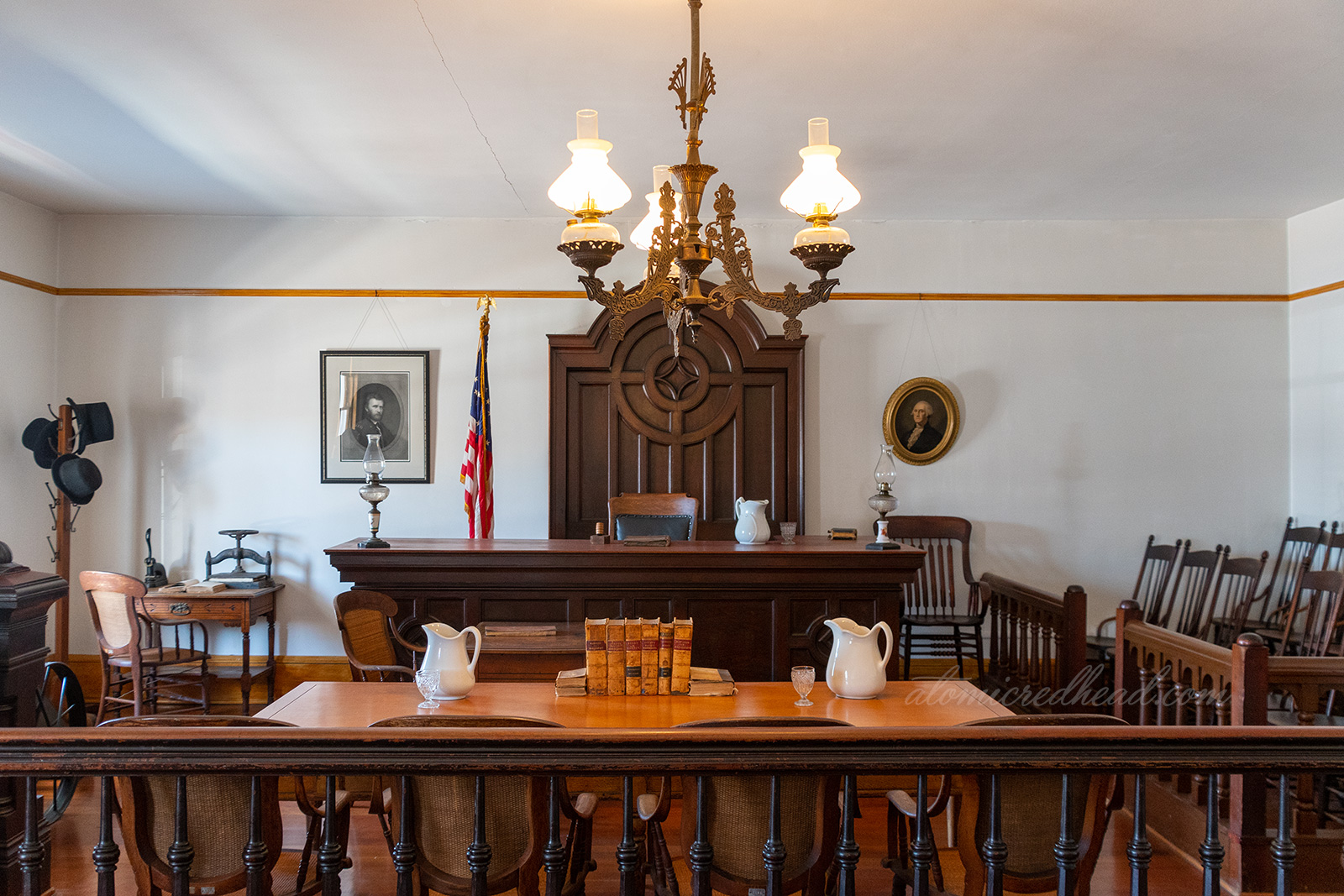 Interior of the courtroom, featuring dark wood seat for the judge, and table for defense and prosecution. An old fashioned brash lamp hangs from the ceiling.
