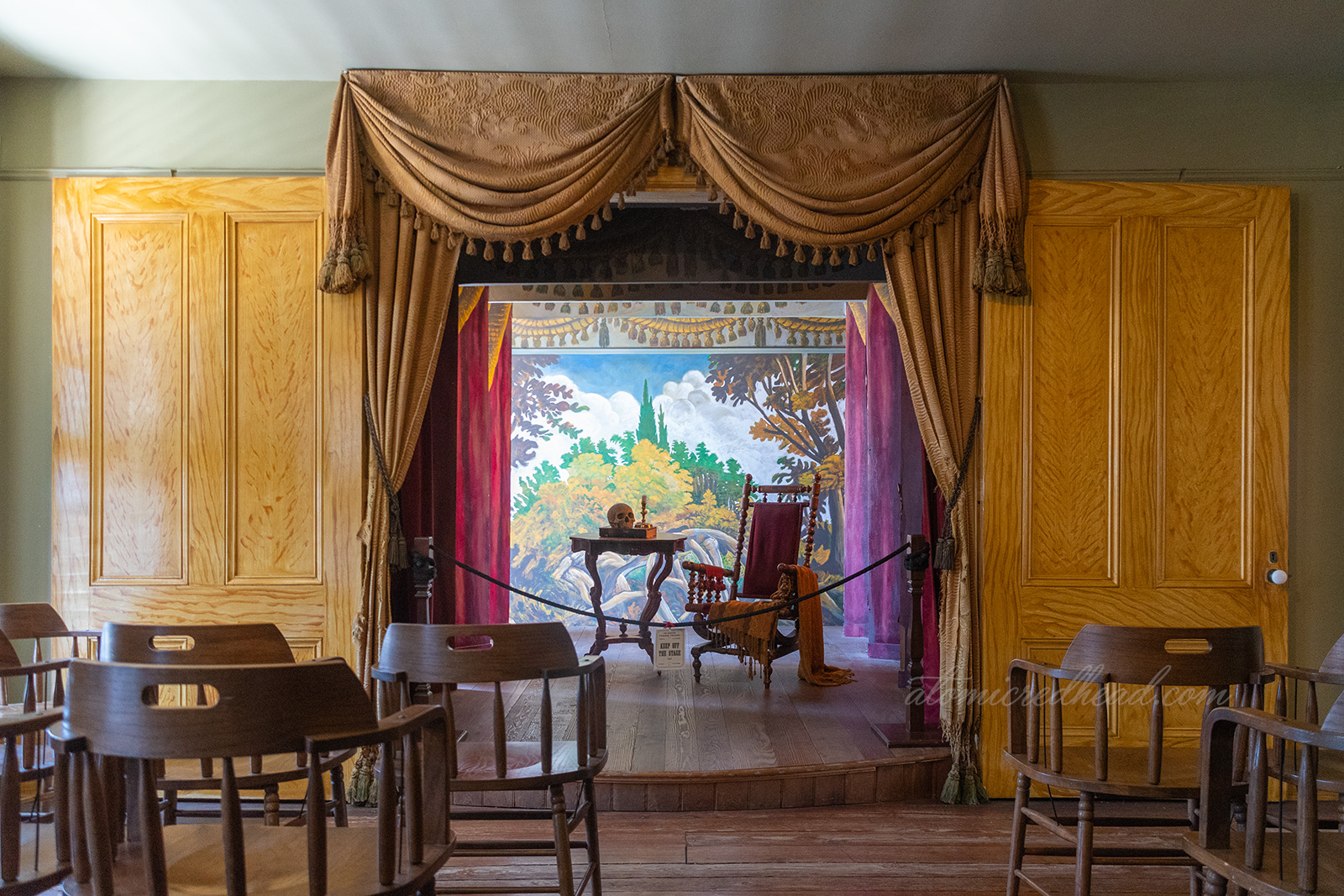 The stage in the room that was the first theatre in San Diego. Gold curtains hang around the stage, where a chair sits with a small table by it, atop the table a skull, candle, and book.