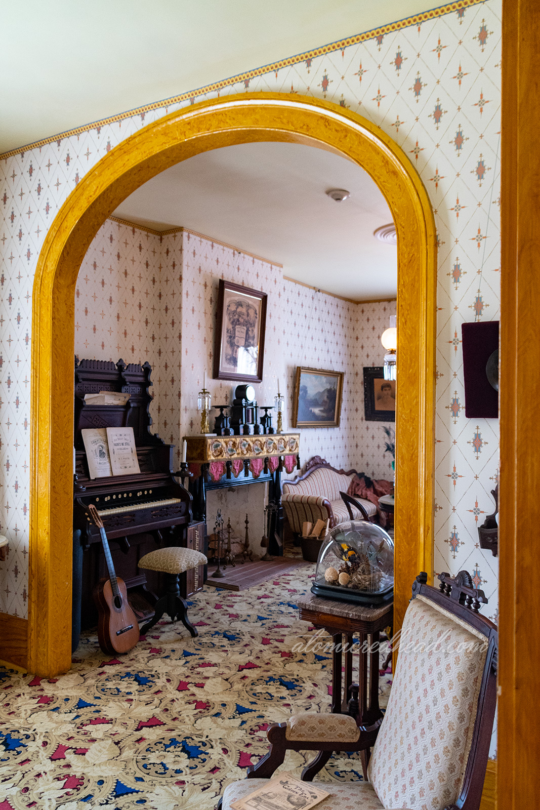 The parlor of the home, with an archway separating two areas. White and gold wallpaper line the walls, a dark wood organ sits agains the wall with a guitar leaning against it, the fireplace is next to it.