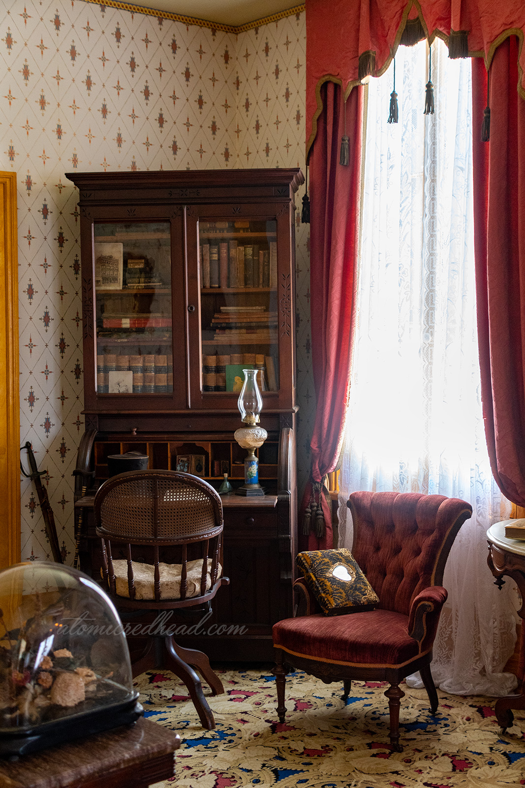Inside the parlor a desk with bookshelf above sits in the corner. Dark red curtains hang from the window.