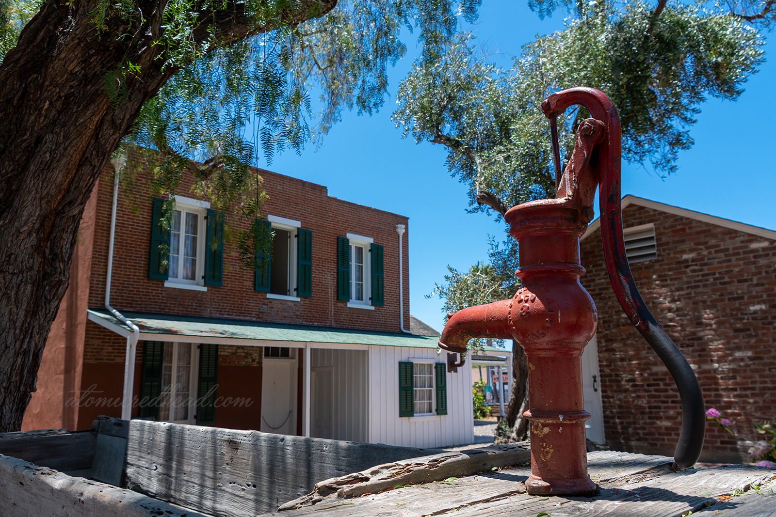 The back of the home, which features exposed brick, windows with green shutters. A red water pump sits in the foreground and is attached to a wooden trough. 